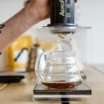 A person uses an AeroPress to brew espresso-style coffee into a glass carafe, illustrating the AeroPress vs French Press comparison.