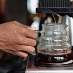 Person brewing coffee with an automatic drip coffee maker, showing a hand holding a glass carafe as coffee drips in.