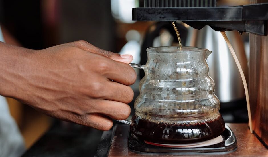 Person brewing coffee with an automatic drip coffee maker, showing a hand holding a glass carafe as coffee drips in.