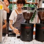 Barista cleaning a coffee grinder with a brush beside two large grinders, illustrating effective grinder cleaning tools.
