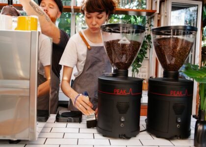 Crop female worker with brush near hipster male partner against professional electric coffee maker and grinders in cafeteria