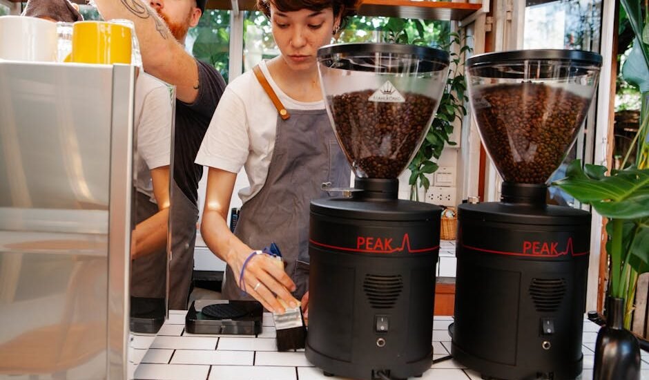 Crop female worker with brush near hipster male partner against professional electric coffee maker and grinders in cafeteria