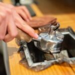 Close-up of a hand knocking used espresso grounds from a portafilter into a metal coffee knock box on a wooden counter.