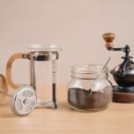 French press coffee gear including a manual grinder, glass jar of grounds, and empty press, illustrating grinder choices.
