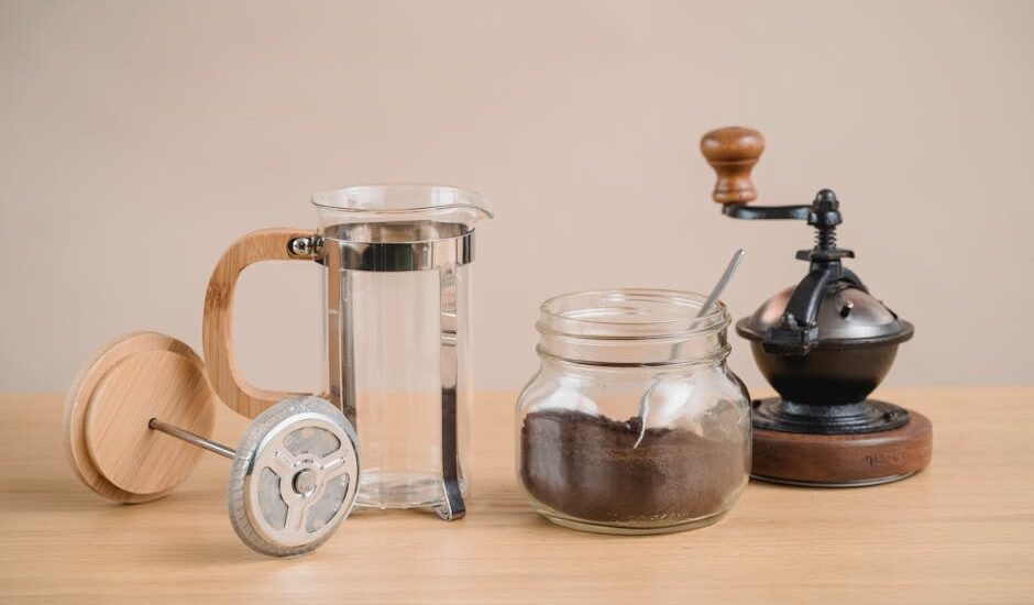 French press coffee gear including a manual grinder, glass jar of grounds, and empty press, illustrating grinder choices.