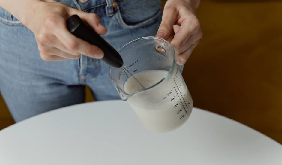 Person using a handheld milk frother to foam milk in a glass measuring cup, illustrating latte preparation for a milk frother guide