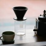 From above of manual coffee grinder with pour over coffeemaker placed in wooden table with ceramic cup and glass jug with roasted beans in morning