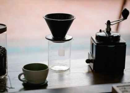 From above of manual coffee grinder with pour over coffeemaker placed in wooden table with ceramic cup and glass jug with roasted beans in morning