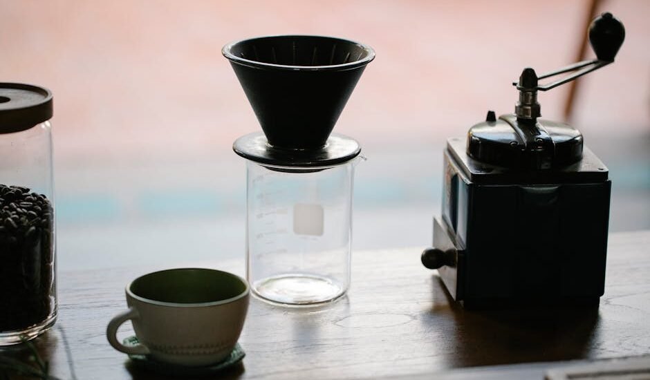 From above of manual coffee grinder with pour over coffeemaker placed in wooden table with ceramic cup and glass jug with roasted beans in morning