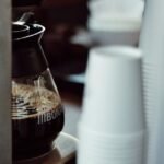 Glass carafe filled with brewed coffee in a drip coffee maker, with stacked disposable cups nearby, illustrating descaling steps.