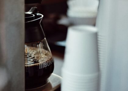 Glass carafe filled with brewed coffee in a drip coffee maker, with stacked disposable cups nearby, illustrating descaling steps.