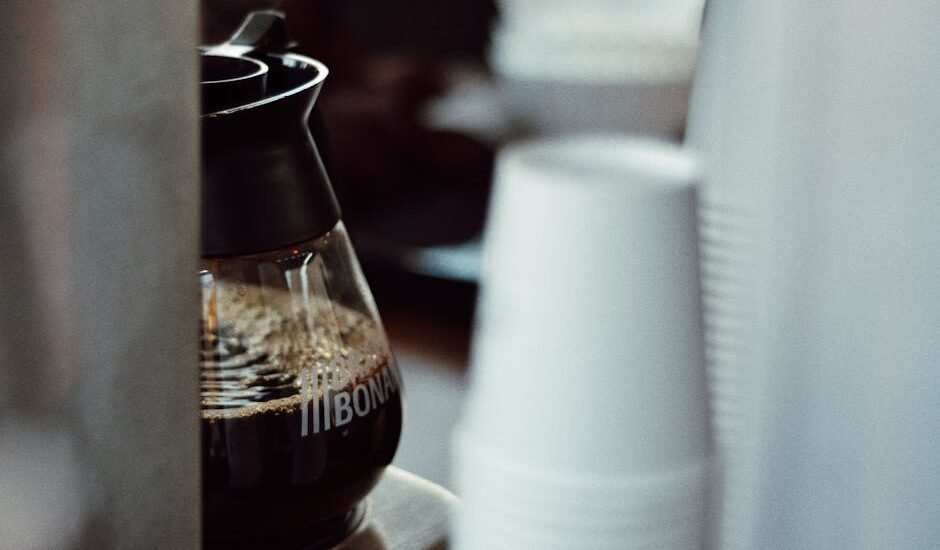 Glass carafe filled with brewed coffee in a drip coffee maker, with stacked disposable cups nearby, illustrating descaling steps.