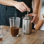 Person frothing milk using a manual plunger frother next to a glass of coffee, illustrating home milk frothing methods.