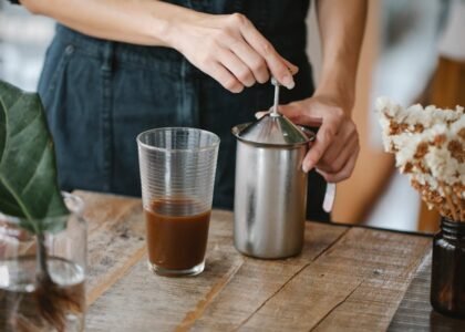 Person frothing milk using a manual plunger frother next to a glass of coffee, illustrating home milk frothing methods.