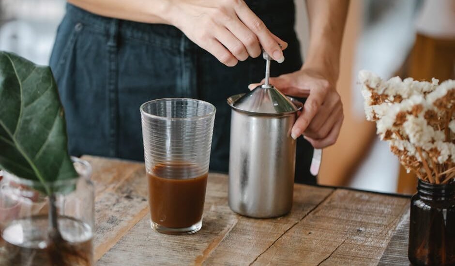 Person frothing milk using a manual plunger frother next to a glass of coffee, illustrating home milk frothing methods.