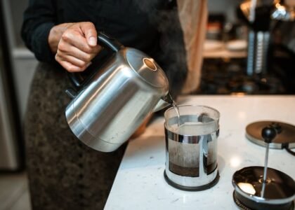 Person pouring hot water from a kettle into a French press coffee maker on a kitchen counter, illustrating home brewing.