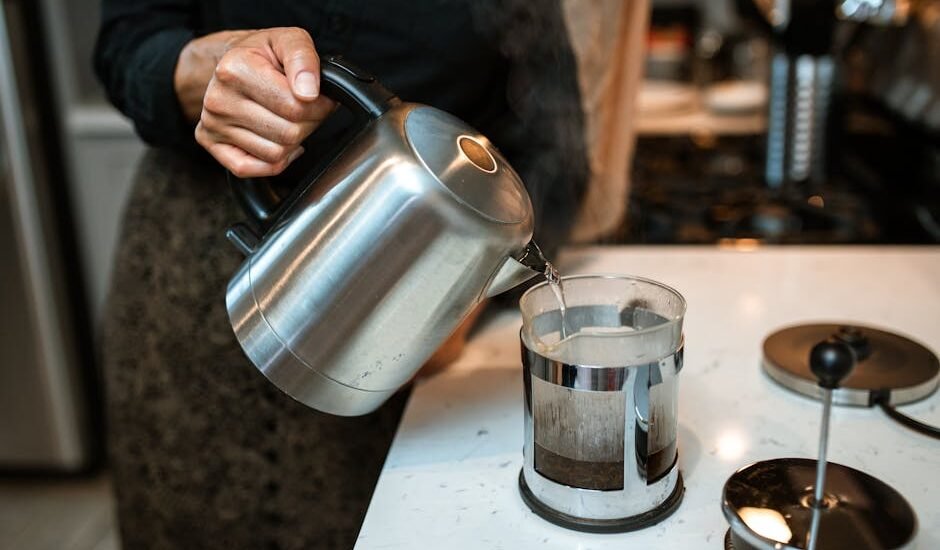 Person pouring hot water from a kettle into a French press coffee maker on a kitchen counter, illustrating home brewing.