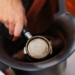 A hand knocks used espresso grounds from a portafilter into a coffee knockout box, illustrating its practical use.