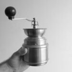 A hand holding a manual conical burr grinder, relevant for drip coffee brewing.