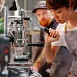 A person operating a manual espresso lever machine while another observes, illustrating the article on manual espresso levers.