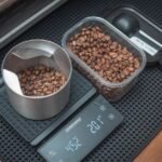 A portable coffee scale displays weight while measuring roasted coffee beans in a bowl and container.