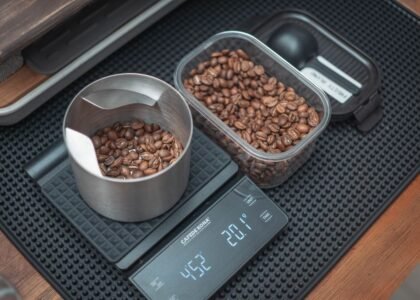 A portable coffee scale displays weight while measuring roasted coffee beans in a bowl and container.
