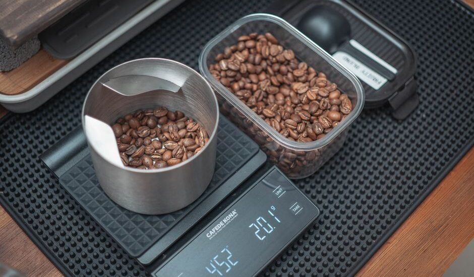 A portable coffee scale displays weight while measuring roasted coffee beans in a bowl and container.