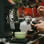 A person holds a portafilter near a coffee dosing cup while preparing espresso.