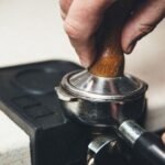 A hand pressing down on an espresso tamper with a wooden handle on a portafilter.