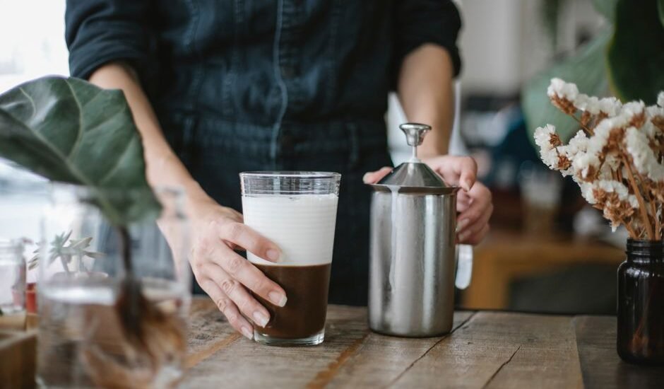 A person holding a glass of frothed milk next to a manual milk frother, illustrating how it works.