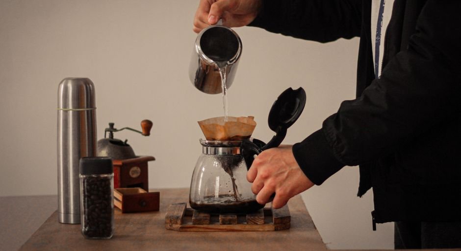 A person pours hot water over a coffee filter in a brewing setup, illustrating how to pre-wet a coffee filter.