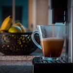 A single serve drip coffee maker pouring coffee into a clear mug, with fruit in the background, illustrating home brewing.