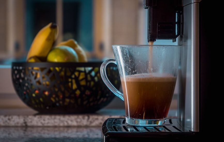 A single serve drip coffee maker pouring coffee into a clear mug, with fruit in the background, illustrating home brewing.